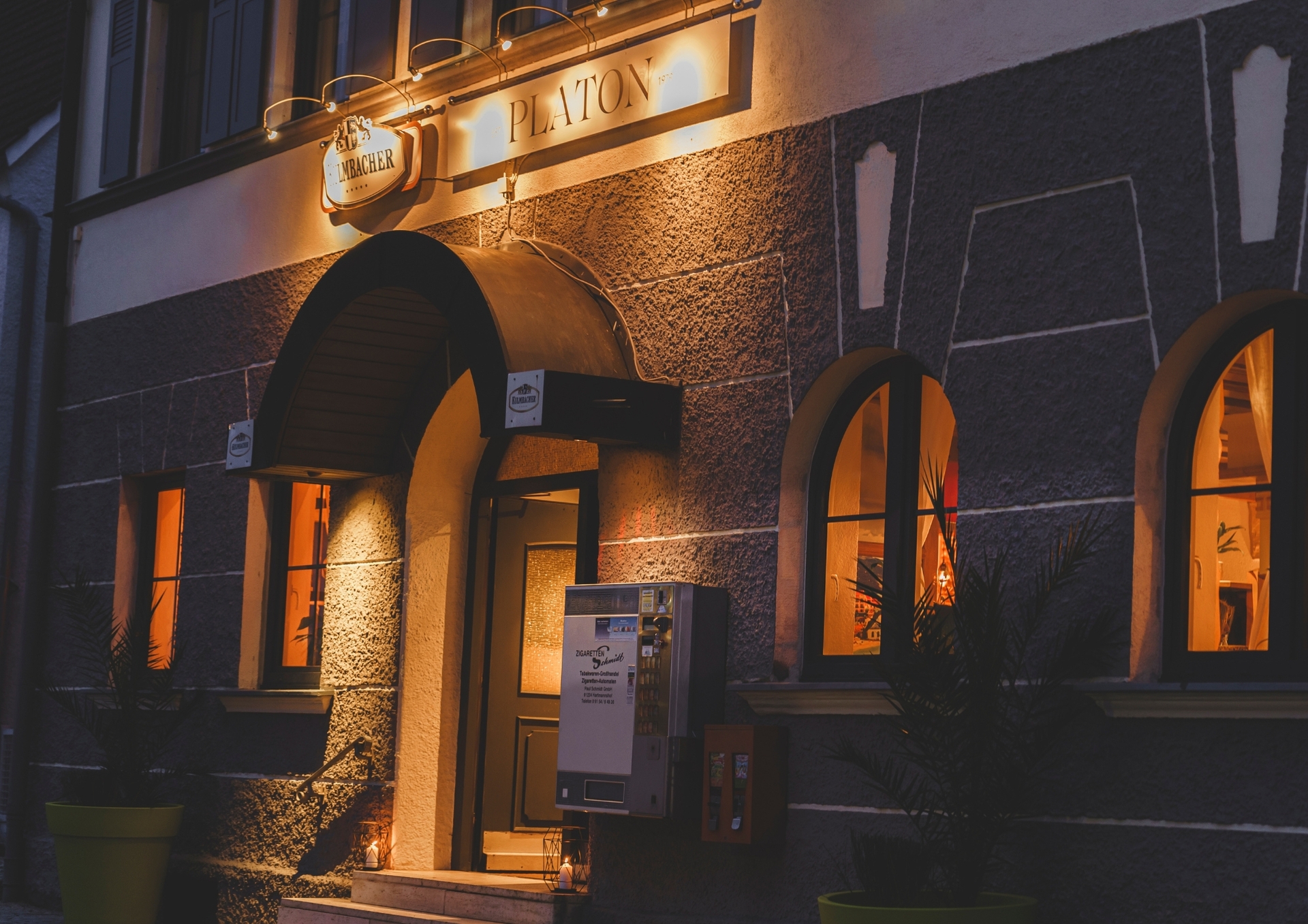Abendansicht des Restaurants Platon mit beleuchtetem Schild über dem gewölbten Eingang und warm beleuchteten Fenstern, die eine gemütliche Atmosphäre in das Gebäude mit Steinfront einladen.