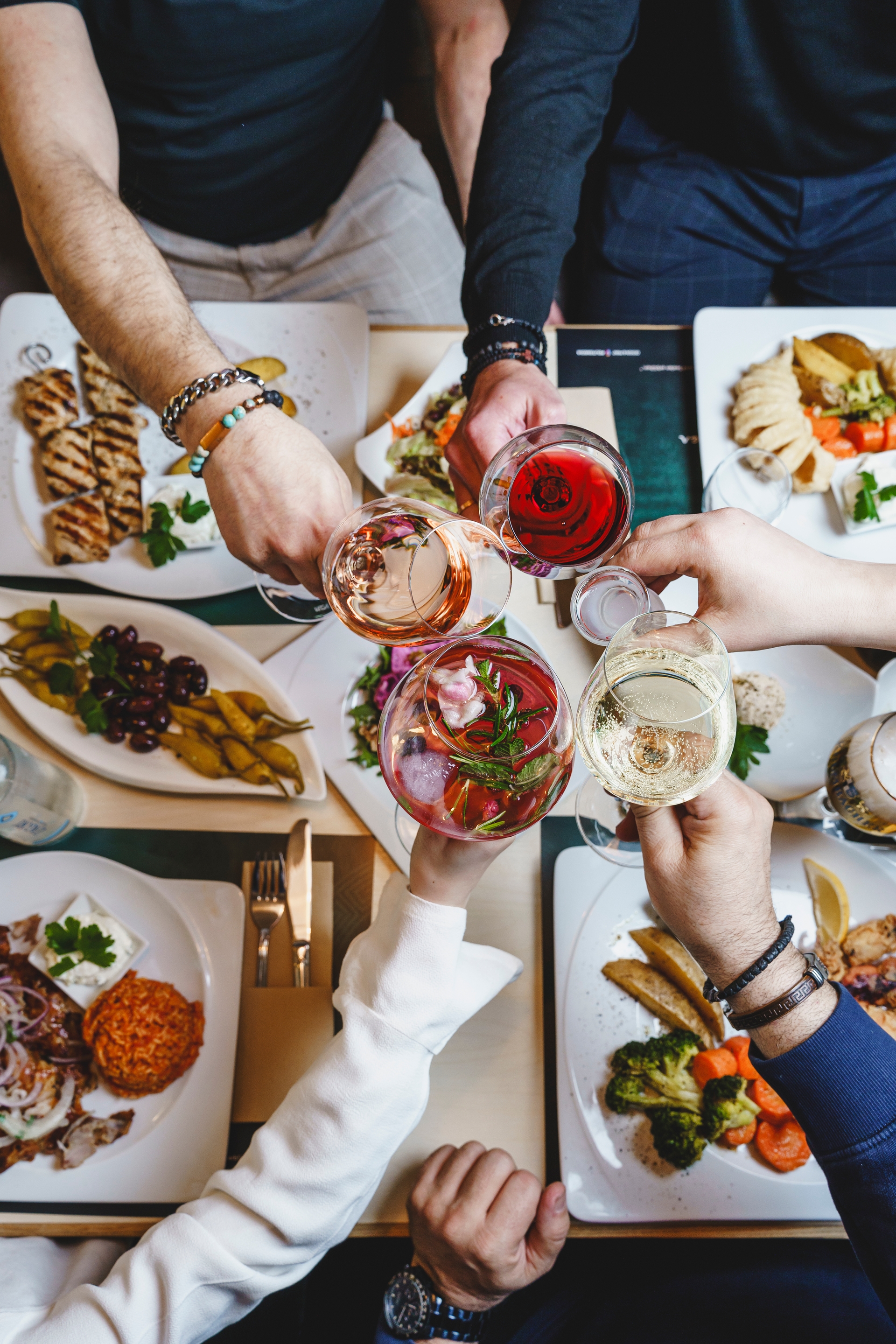 Menschen beim Essen im Restaurant, die Gläser mit Wein über einen Tisch voller Gerichte heben, darunter gegrilltes Hähnchen, Gemüse und traditionelle griechische Meze.