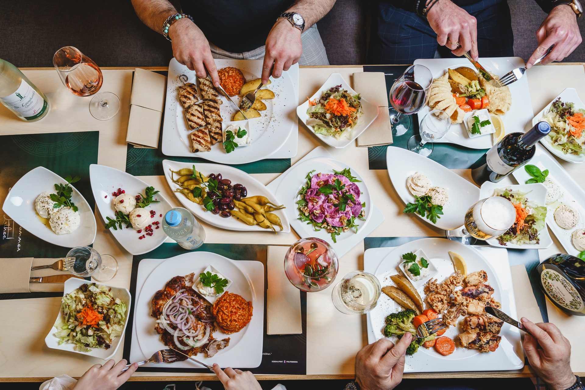 Menschen beim Essen im Restaurant, die Gläser mit Wein über einen Tisch voller Gerichte heben, darunter gegrilltes Hähnchen, Gemüse und traditionelle griechische Meze.